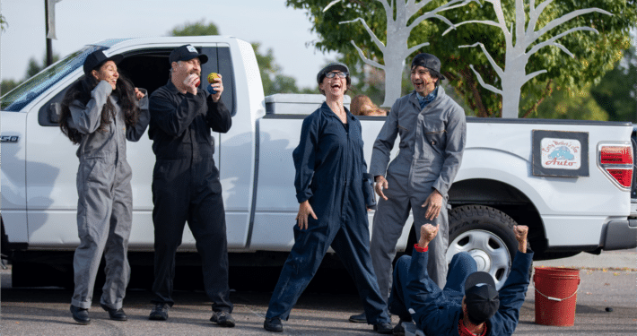 In front of a white pickup truck, ensemble of actors in carpenter suits cheer on actress in a newsboy cap