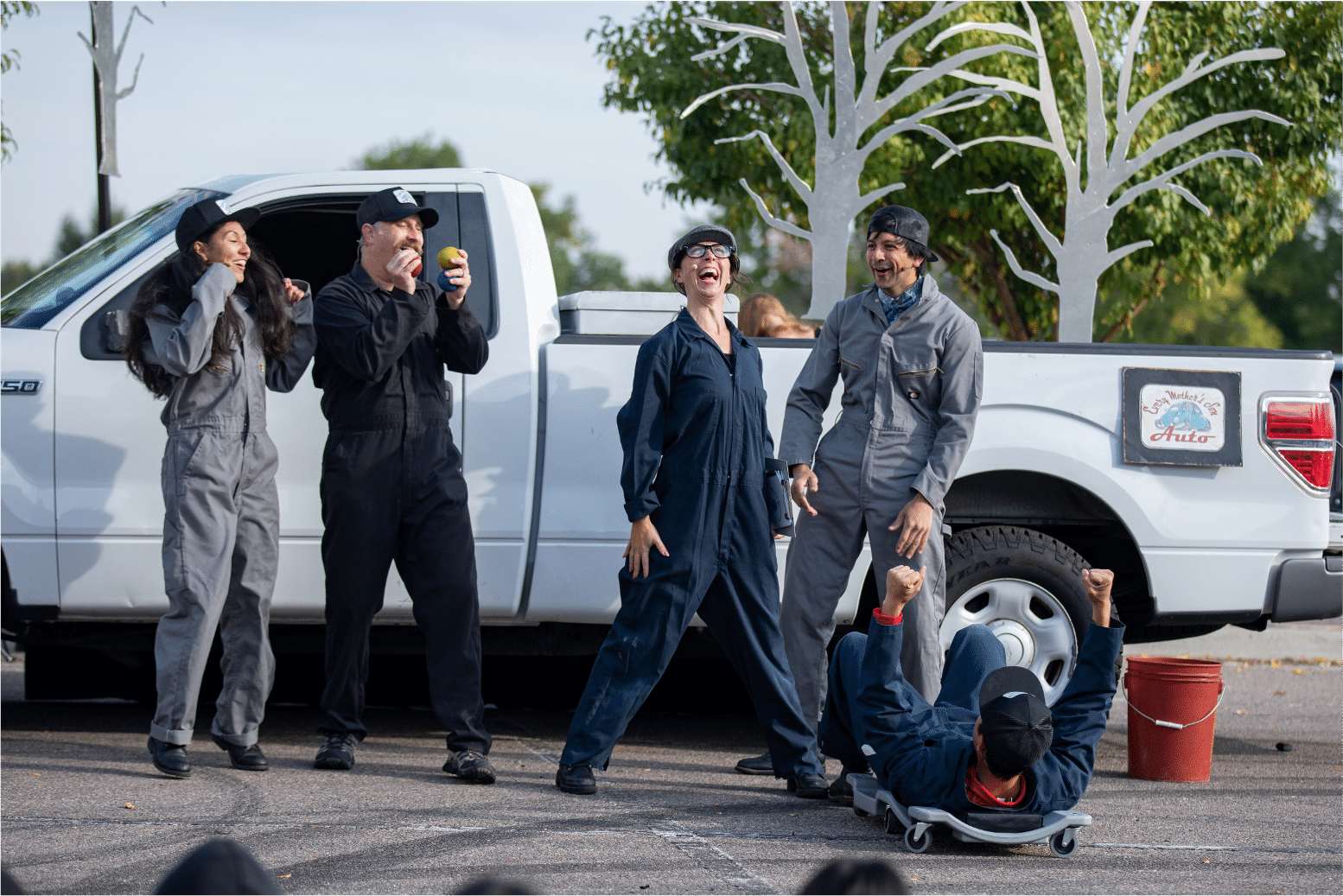 In front of a white pickup truck, ensemble of actors in carpenter suits cheer on actress in a newsboy cap