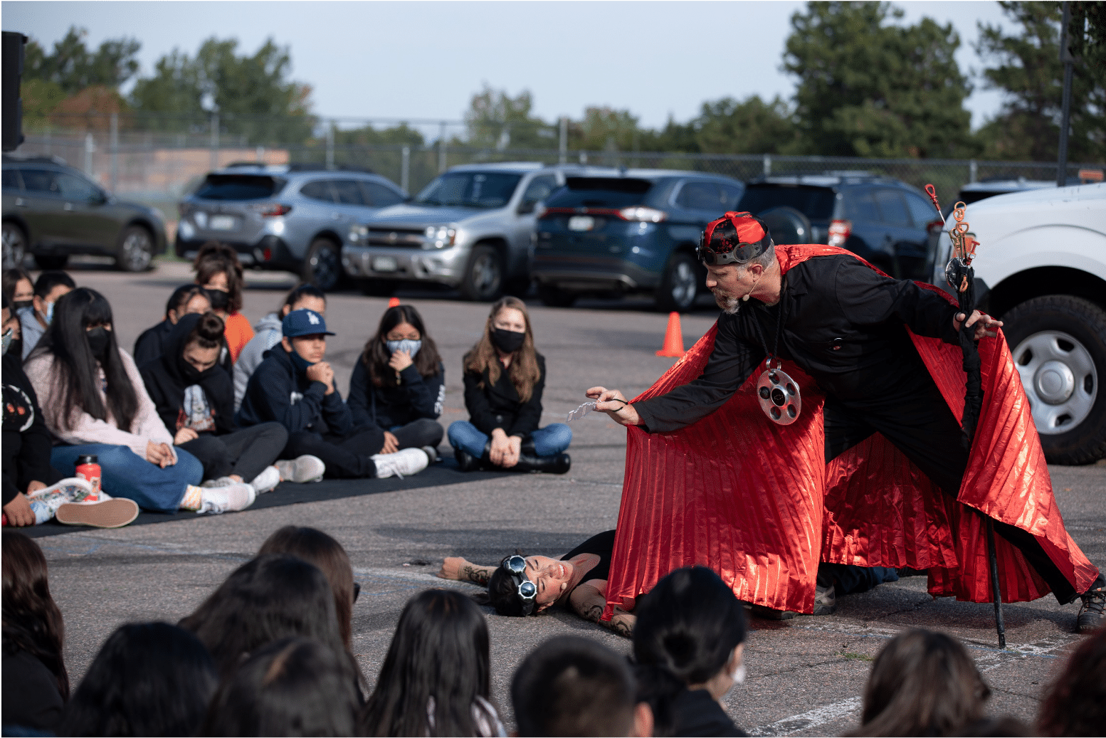 Actor with tattoos lays unconscious as a man in an elegant ruby red cape with a fancy cane hover over them