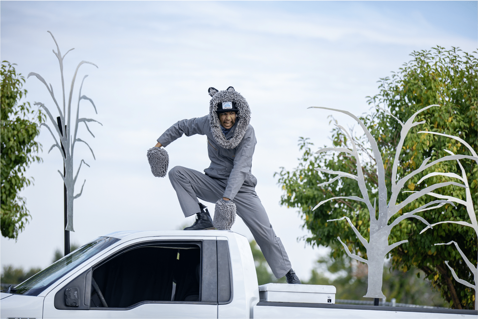 Man in a grey wolf costume perches on top of a white pickup truck snarling down below