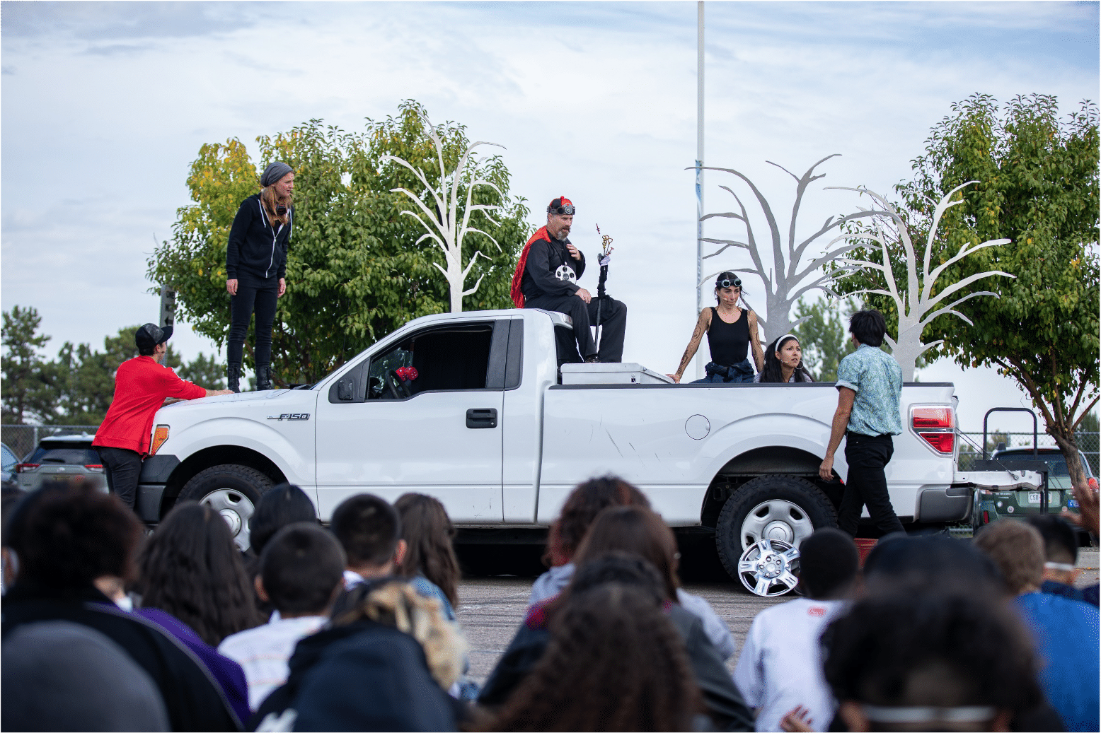 Ensemble of actors sit at various spots on a white pickup truck as they stare at a man walking toward them in a blue shirt