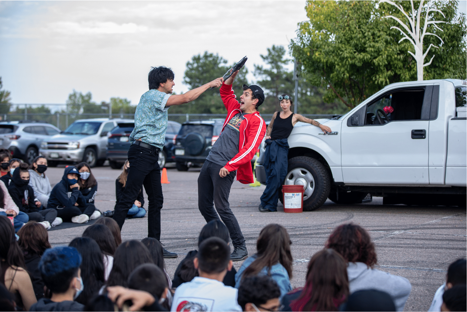 Man in blue shirt and a man in a red jacket fight over a dark object during a production of Shakespeare in The Parking Lot