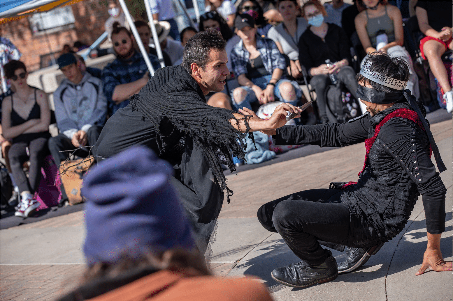 Actor in black tatters holds dagger towards a blindfolded actor in a silver crown and red sash on the ground in fear