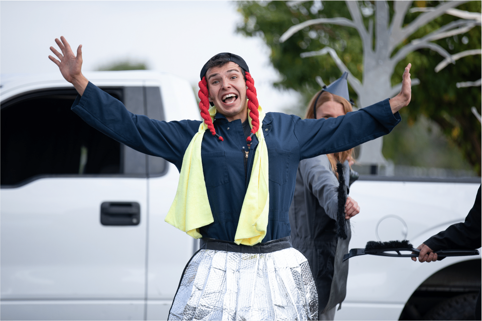 Man smiles at audience with arms open crossdresses as a woman with red braided pigtails, a yellow scarf, and a silver skirt