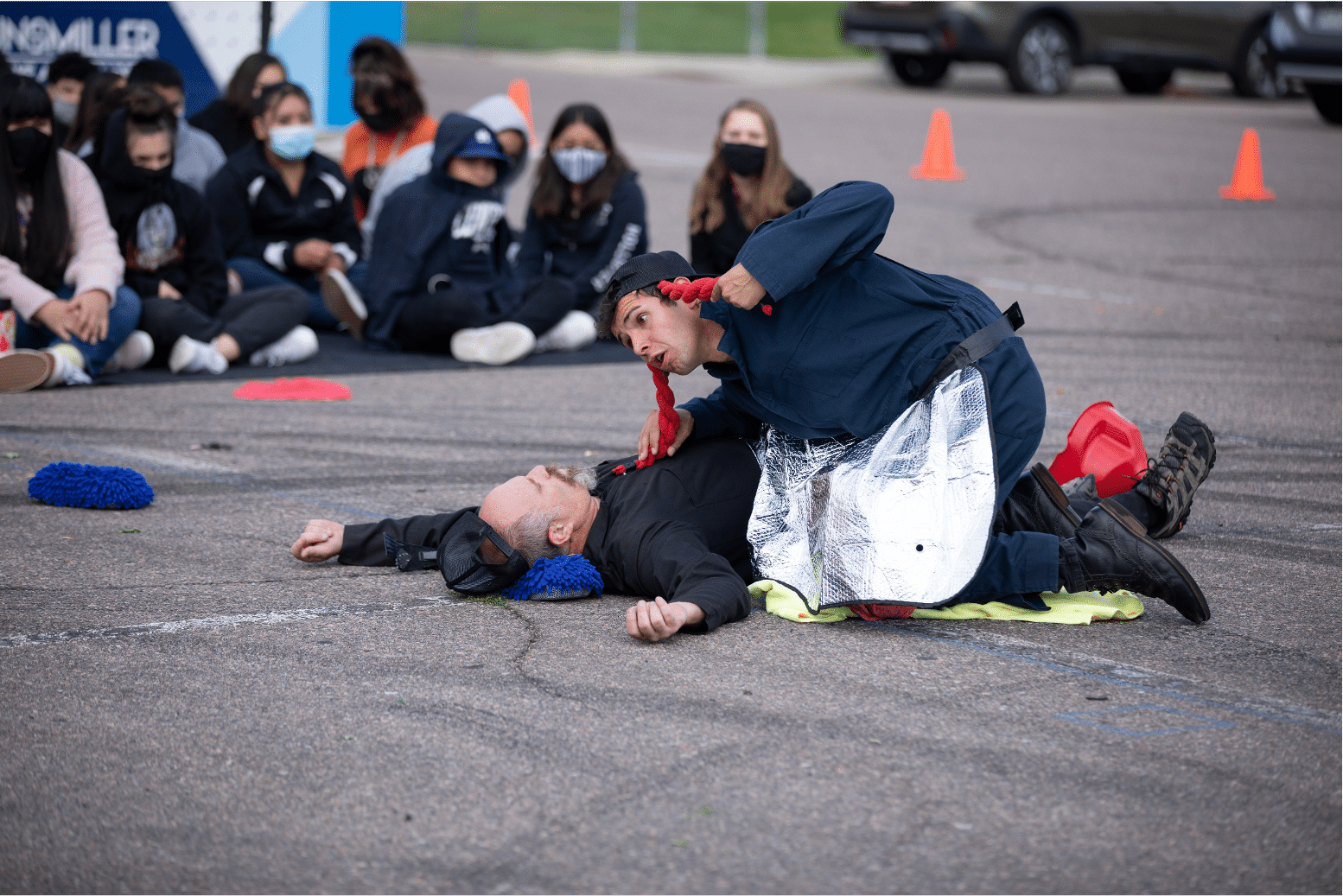 Actor lays unconscious while a crossdressing man hovers over and uses his red braided pigtail as a sort of stethoscope