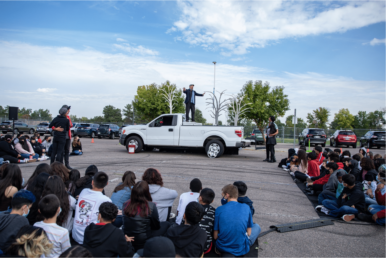 Man stands on bed of a white pickup truck shouting to the young audience as two couples embrace each other on opposite ends