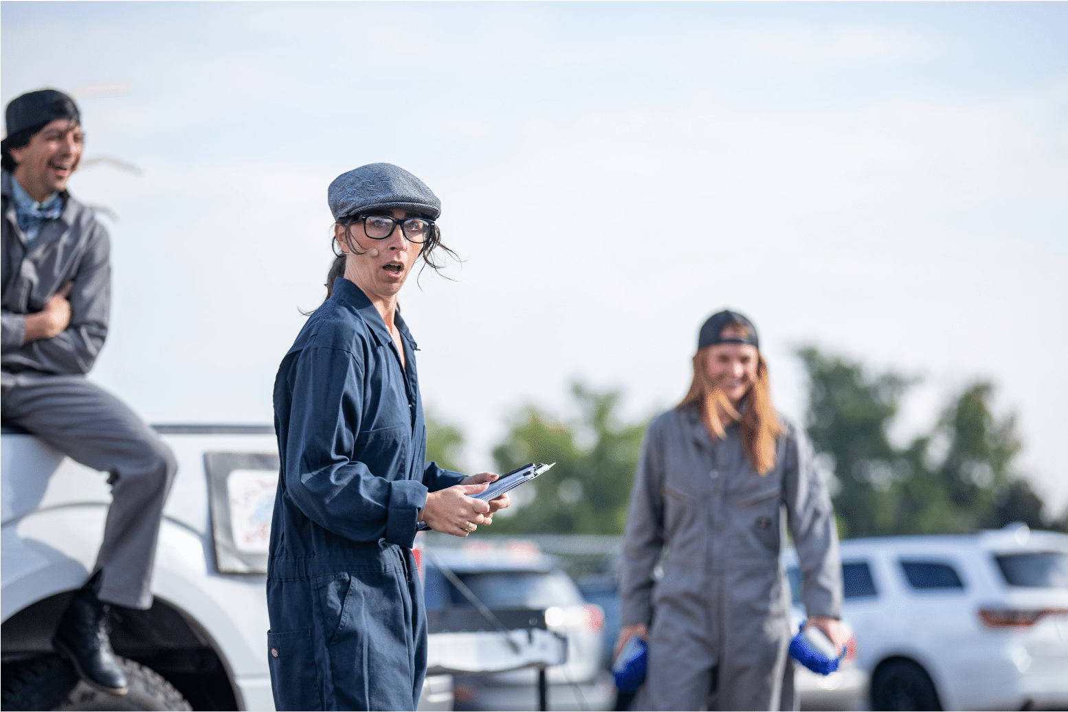 Actor in navy carpenter suit stares out in shock and disbelief. Blurred behind, two actors laugh and smile at the ground