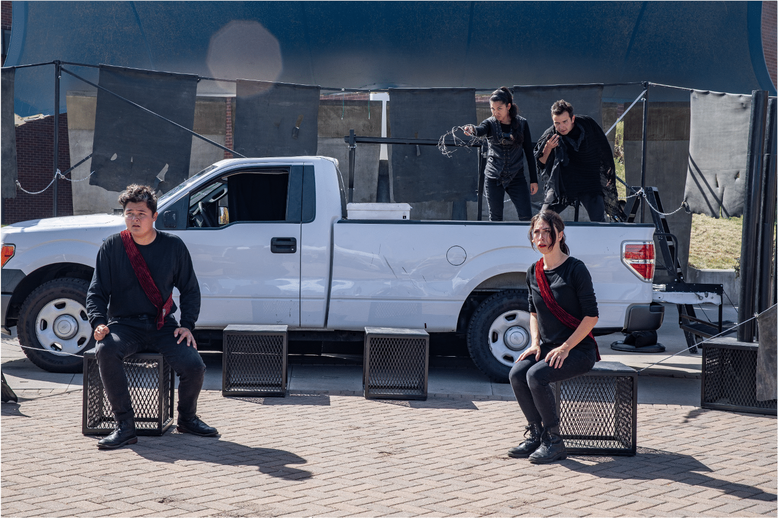 Two actors on blocks stare in shock outwards. Behind them, two actors in bed of a pickup truck point and stare in disgust.