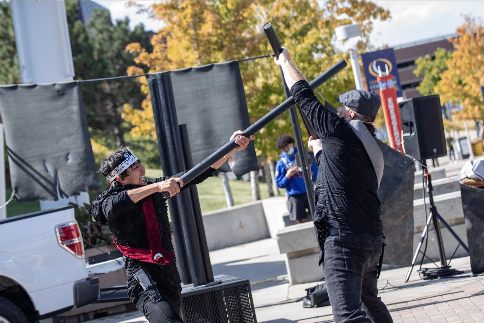 Two men fight with large black pipes. One in red sash and silver crown and one in a silver sash and newsboy cap