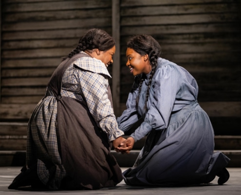 Two women kneel, facing one another with their hands clasped. They are both smiling brightly.