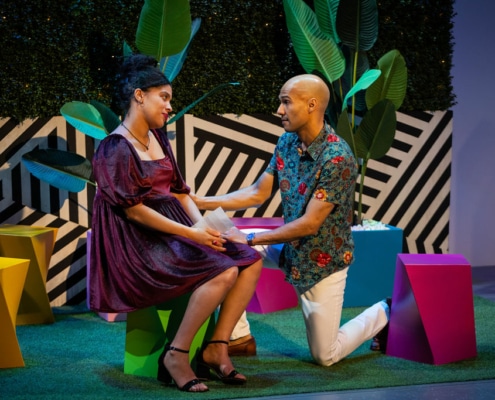 Danielle Alonzo sits on a stool in a colorful courtyard while Luis Vega kneels in front of her holding her hand.