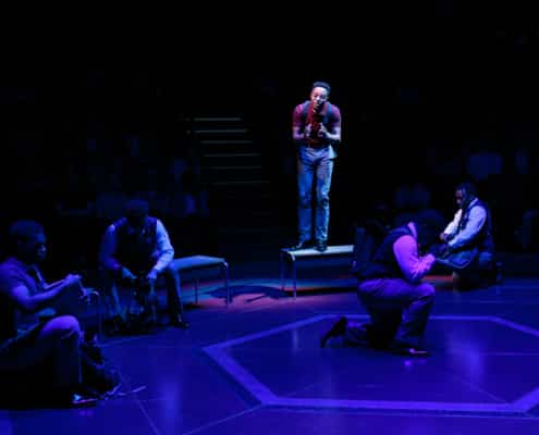 Four school uniformed males sit in a dim lighted setting while the spotlight shines on a male standing on a bench