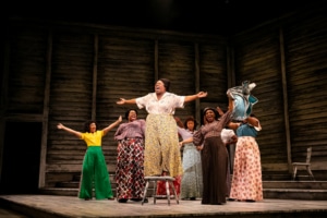 Several women sing onstage, clustered together with their arms outstretched. The woman in front is standing on a chair as she belts out a note.