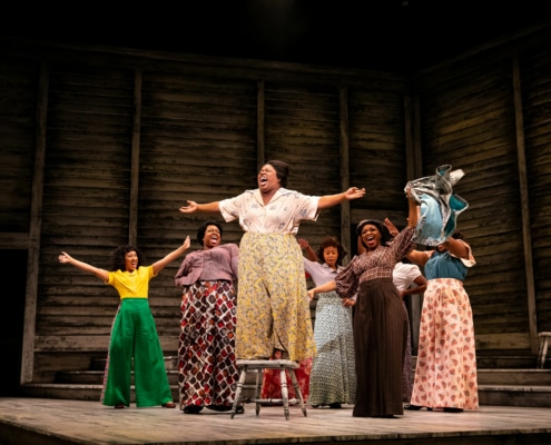 Several women sing onstage, clustered together with their arms outstretched. The woman in front is standing on a chair as she belts out a note.