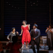 A woman sings while standing on a chair. She's wearing a glittering red dress. People clap and dance around her.