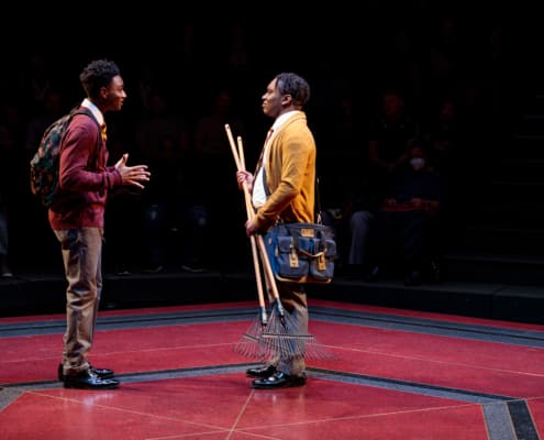 Uniformed boy on the left is speaking to his school mate on the right who is holding two rakes