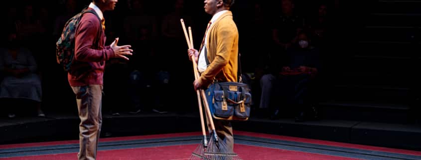 Uniformed boy on the left is speaking to his school mate on the right who is holding two rakes