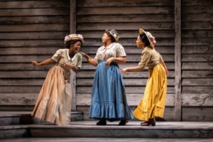 Three women wearing decorative hats and long, colorful skirts laugh and talk with one another.