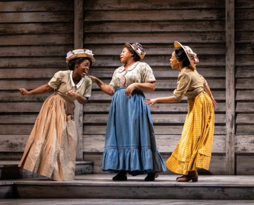 Three women wearing decorative hats and long, colorful skirts laugh and talk with one another.