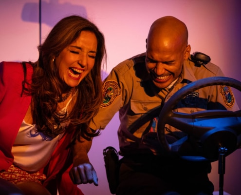 Maggie Bofill and Luis Vega laugh together in front of a raised steering wheel that is meant to portray a police car. Red and blue lights fill the background.