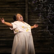 A woman stands with her arms outstretched, belting a song onstage.