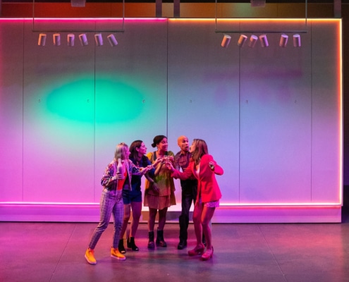 Olivia Hebert, Stephanie Machado, Danielle Alonzo, Luis Vega, and Maggie Bofill cheers shot glasses of coquito in the middle of the stage while colorful lights fill the background.