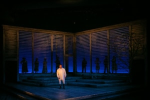 A man stands alone onstage with the shadows of of several people standing behind him outlined in the set backdrop.