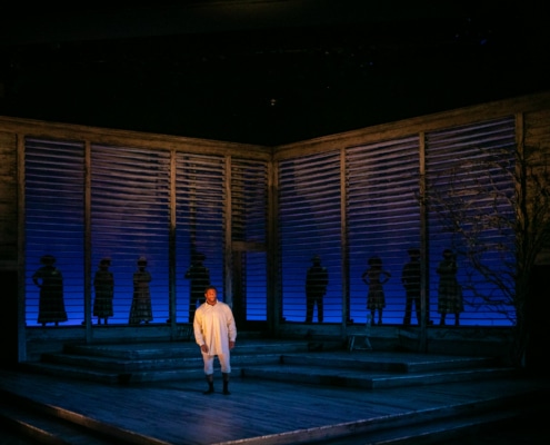 A man stands alone onstage with the shadows of of several people standing behind him outlined in the set backdrop.