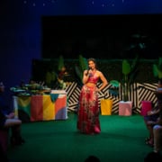 Maggie Bofill holds one hand on her hip and a microphone in the other while standing in the middle of a colorful courtyard. There is an elaborate spread of food on a table behind her, and Stephanie Machado, Danielle Alonzo, and Luis Vega sit watching her.