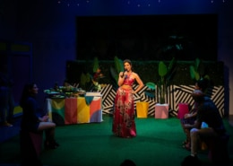 Maggie Bofill holds one hand on her hip and a microphone in the other while standing in the middle of a colorful courtyard. There is an elaborate spread of food on a table behind her, and Stephanie Machado, Danielle Alonzo, and Luis Vega sit watching her.