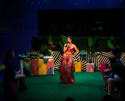 Maggie Bofill holds one hand on her hip and a microphone in the other while standing in the middle of a colorful courtyard. There is an elaborate spread of food on a table behind her, and Stephanie Machado, Danielle Alonzo, and Luis Vega sit watching her.