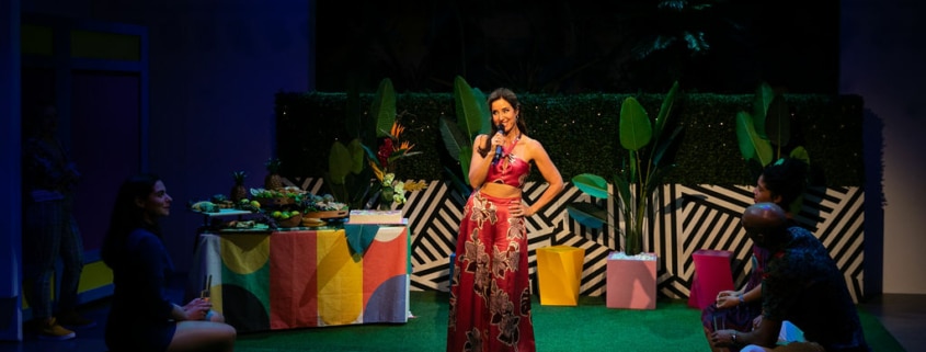 Maggie Bofill holds one hand on her hip and a microphone in the other while standing in the middle of a colorful courtyard. There is an elaborate spread of food on a table behind her, and Stephanie Machado, Danielle Alonzo, and Luis Vega sit watching her.