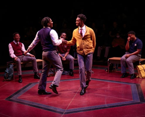 Two males are dancing mid stage in front of three other school mates sitting on the benches behind