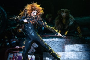 An actor dressed in a black cat costume dances onstage in Cats