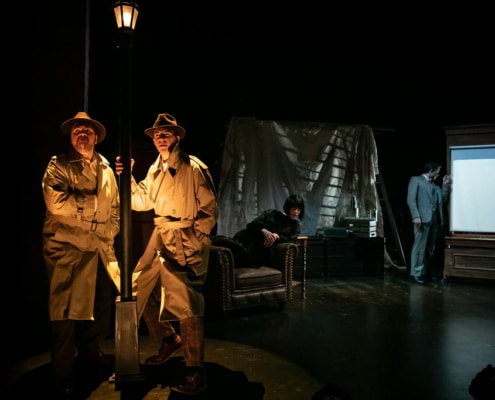 Two mysterious men in trench coats stand under a streetlamp. Annabella Schmidt leans across an armchair and Richard Hannay peers through a window trying not to be seen.