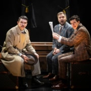Richard Hannay and two traveling salesmen in suits on a train stare out at the audience looking concerned.