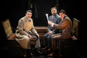 Richard Hannay and two traveling salesmen in suits on a train stare out at the audience looking concerned.