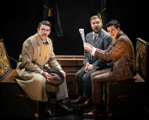 Richard Hannay and two traveling salesmen in suits on a train stare out at the audience looking concerned.