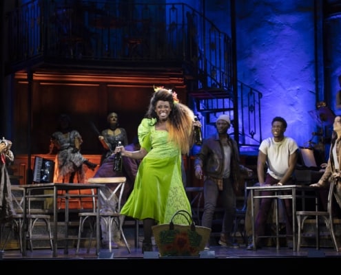 Actress stands mid stage in a bright green dress dancing while cast behind her is looking and listening to her