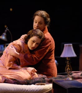 A woman hugs a young woman close while sitting on a bed