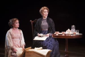 A woman is seated at a table next to a woman sorting through a box