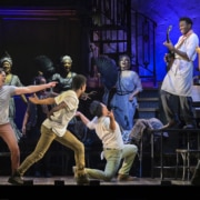 A man standing on a table plays guitar while other cast members react in Hadestown