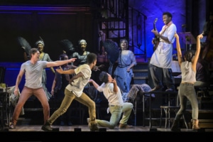 A man standing on a table plays guitar while other cast members react in Hadestown