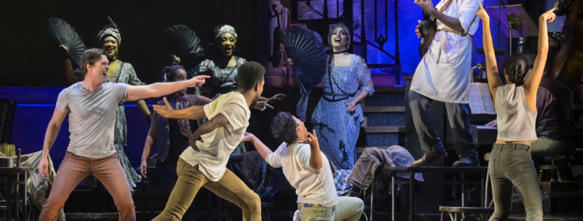 A man standing on a table plays guitar while other cast members react in Hadestown