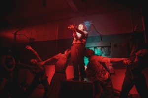 Dancers move around an actor with his hands clasped illuminated in red light