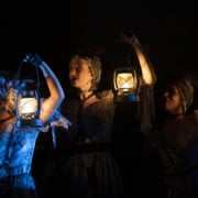 Three women hold lanterns near their faces in Hadestown