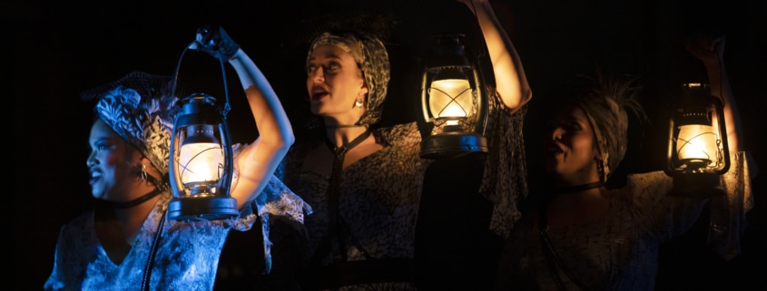 Three women hold lanterns near their faces in Hadestown