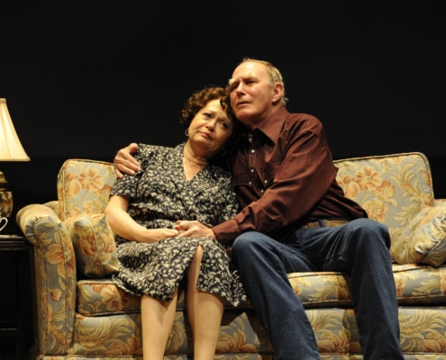 An older man and woman embrace while seated on a couch in Eventide.