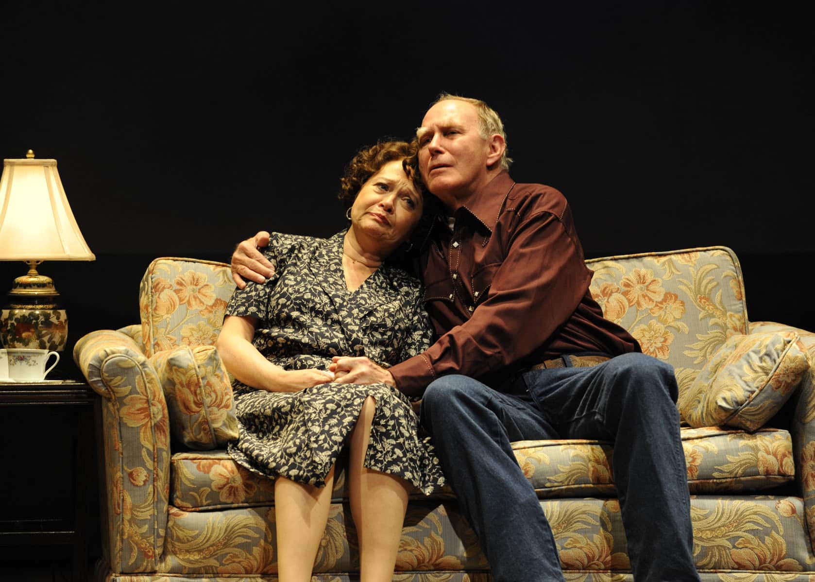 An older man and woman embrace while seated on a couch in Eventide.