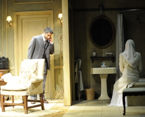 A man stands outside a bathroom where a woman is seated looking at herself in the mirror in Inana.